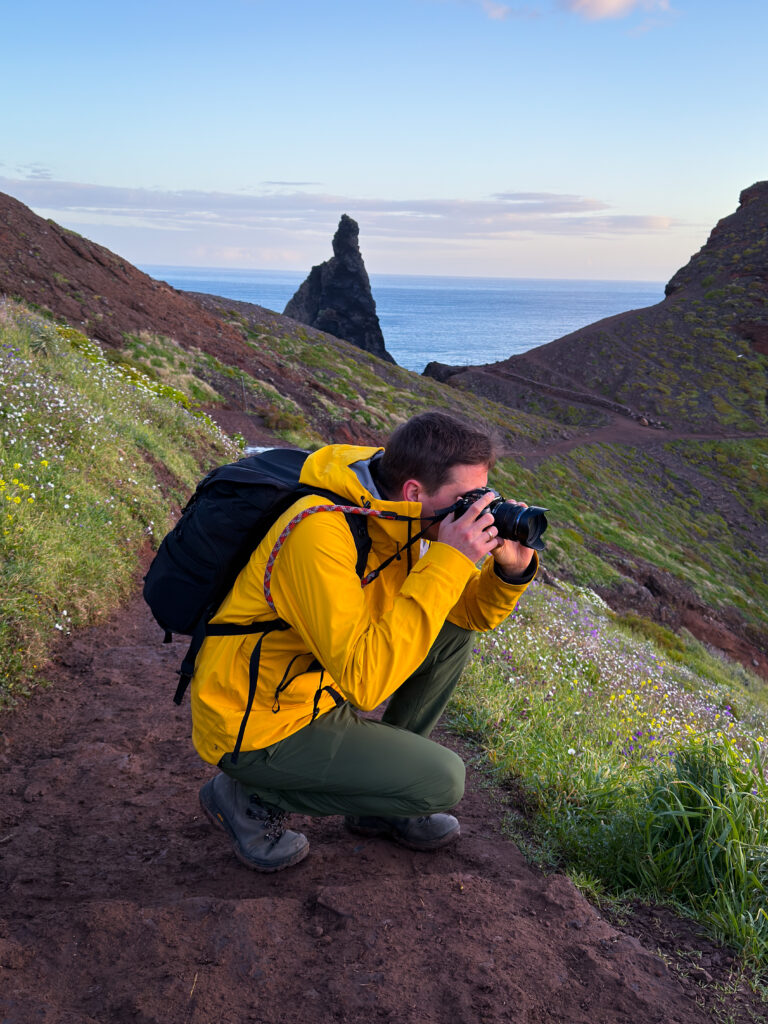 Moritz trägt eine gelbe Regenjacke, eine Kamera und einen schwarzen Kamerarucksack. Er kniet, während er ein Foto aufnimmt. Im Hintergrund grün und braun die zackige Landschaft São Lourenços.