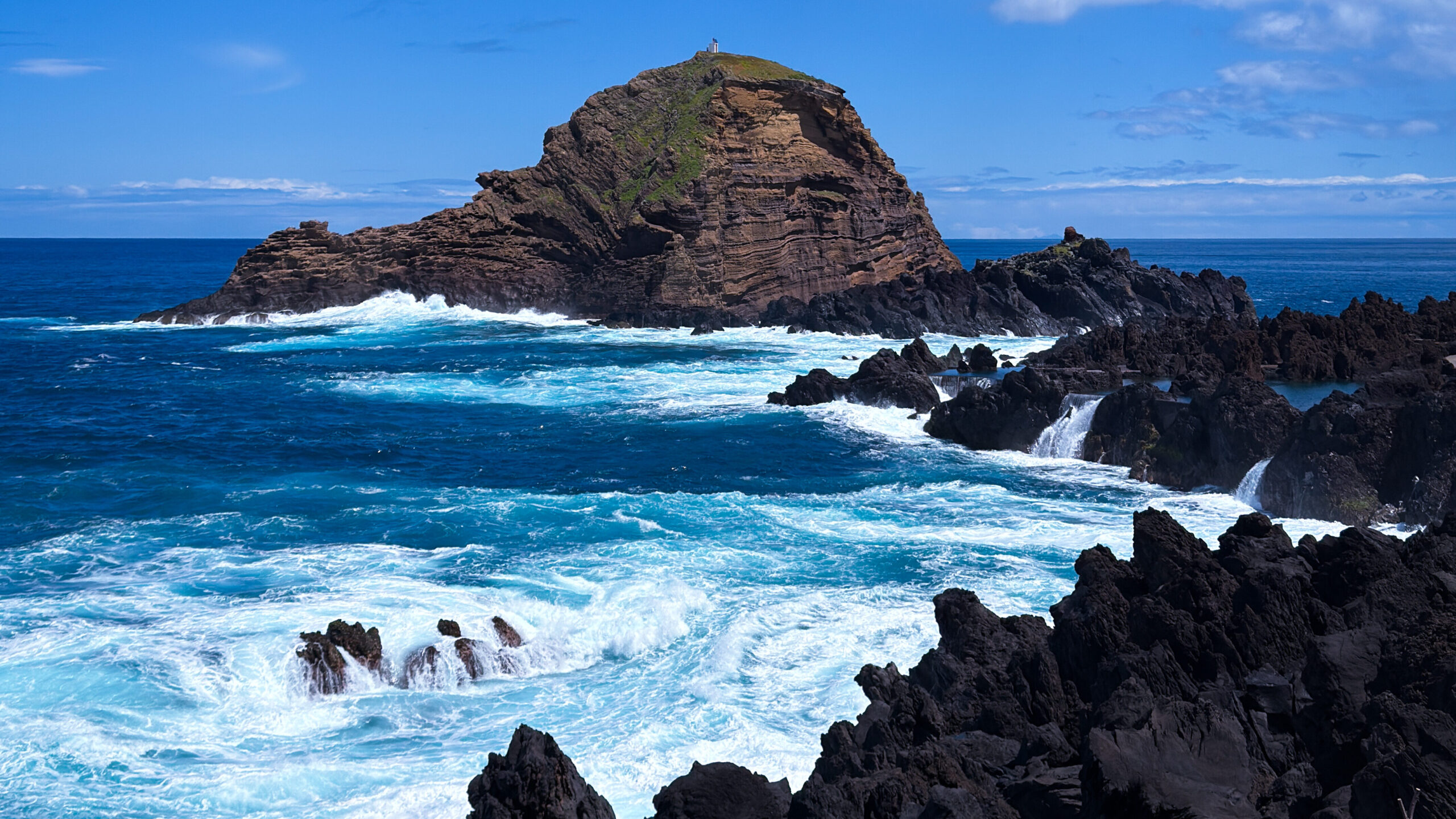 Blick vom Küstenort Porto Moniz auf das azurblaue Meer, auf dem Wellen weiße Schaumkronen schlagen. Im Vorder- und Hintergrund hohe Felsen.