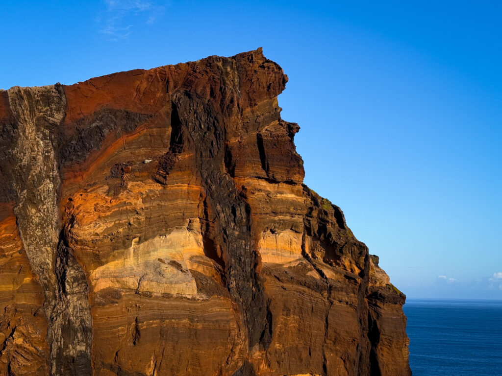 Ein Felsen mit verschiedenen Gesteinsschichten, die sich in unterschiedlichen Farbnuancen zeigen. Dahinter blauer Himmel und blaues Meer.