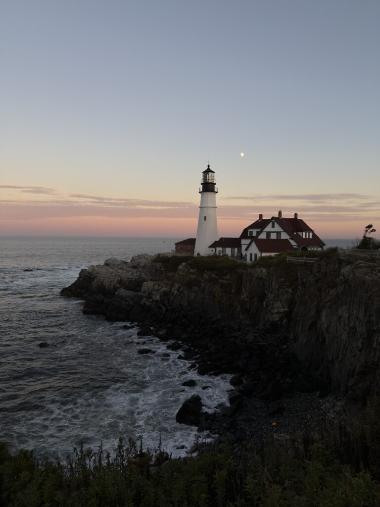 Ein berühmter Leuchtturm im Abendlicht der untergehenden Sonne, der Himmel ist rot orange verfärbt. Neben dem weißen Turm ist der fast volle Mond aufgegangen.