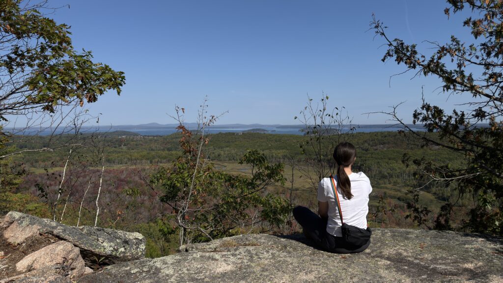 Vanessa sitzt mit dem Rücken zur Kamera an einem Felsen und blickt in die Weite des Acadia Nationalparks