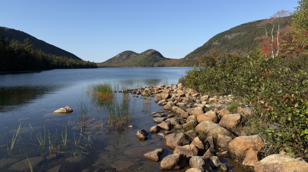 See, im Vordergrund Felsen und bunt gefärbte Bäume.