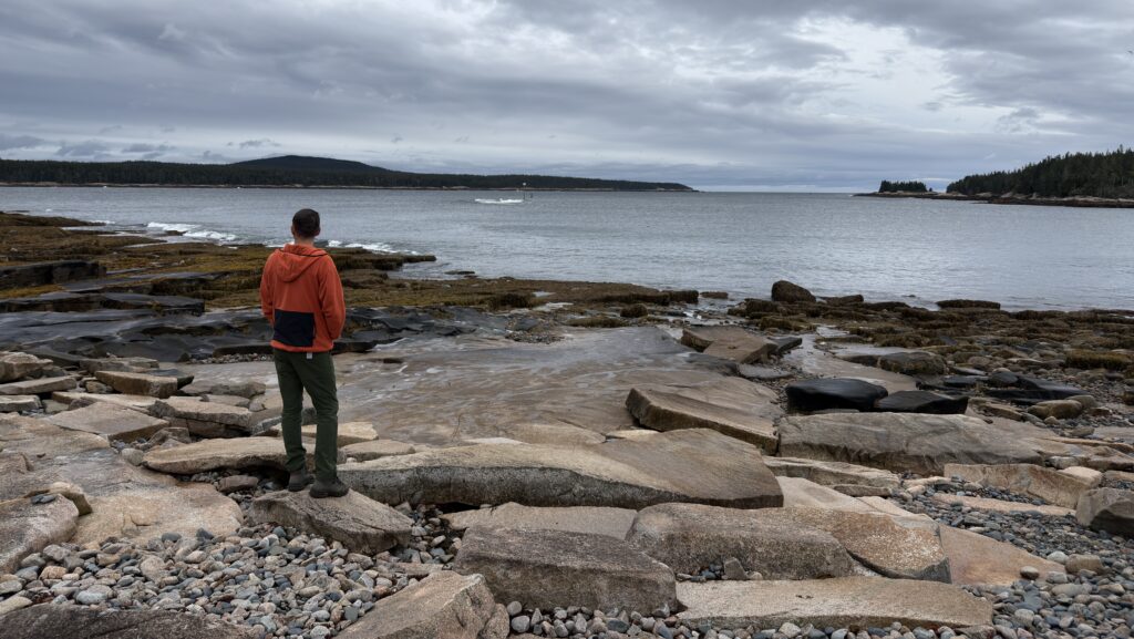 Moritz steht in einem orangenen Pulli am Schoodic Point und blickt auf den Atlantik. 